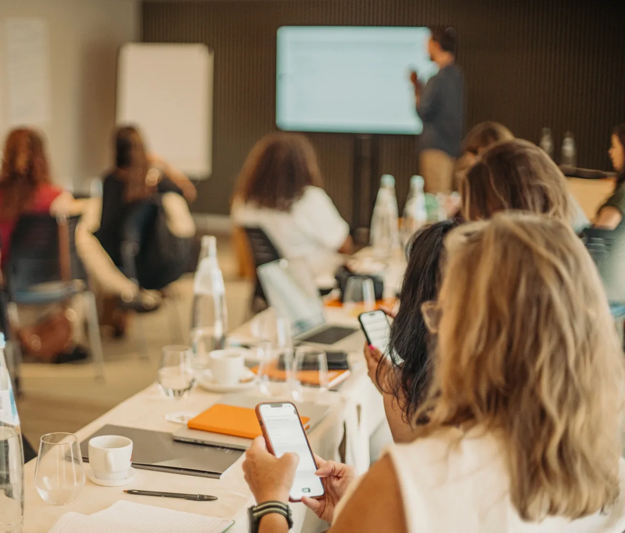 People at a long table during a DDG workshop, two in the foreground looking at their phones while a presenter speaks at a screen behind them.