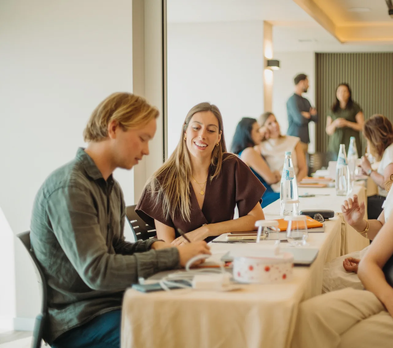Two people at the end of a long table during a DDG workshop, one writing while the other smiles at him, the rest of the room behind them.